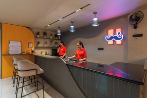 A hotel reception area with a counter, two staff members, a neon sign, and a bulletin board with information. - Moustache Jaipur (Jaipur)
