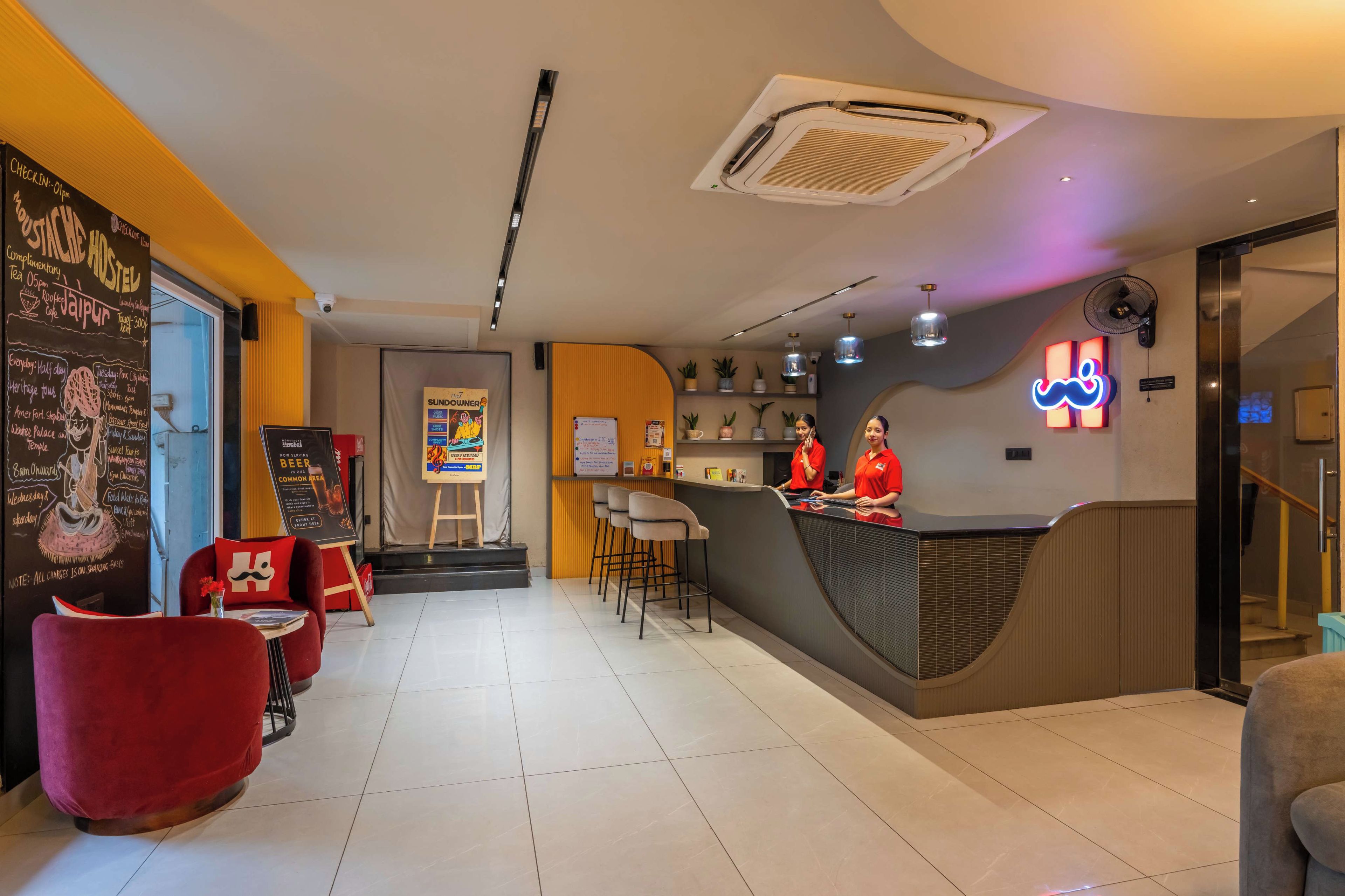 Un interno moderno di un ostello, con una reception, un'area salotto e una lavagna con gli eventi in programma.