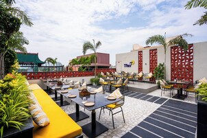 Un área de comedor al aire libre con mesas y sillas, rodeada de follaje y un edificio con un letrero que dice 'The7'.