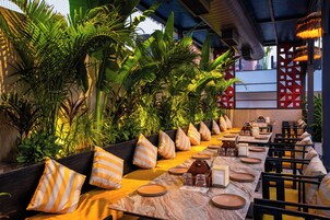 Uma longa mesa de jantar com almofadas listradas, rodeada por plantas tropicais exuberantes, com pratos, copos e talheres.