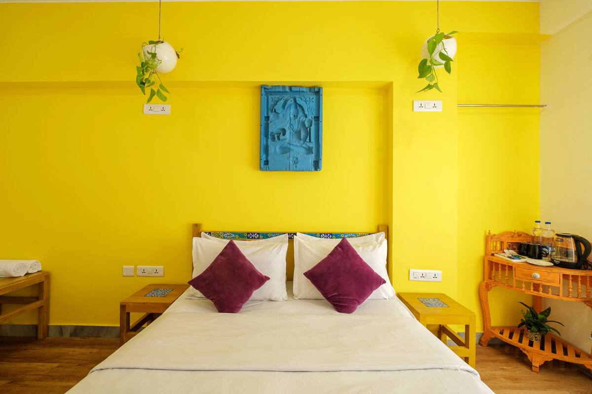 A bright yellow room with a bed, two purple pillows, a blue artwork on the wall, and a wooden side table with a plant.