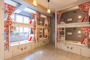 Quarto de hotel com beliches, cada um com cortina, janela e ventilador.