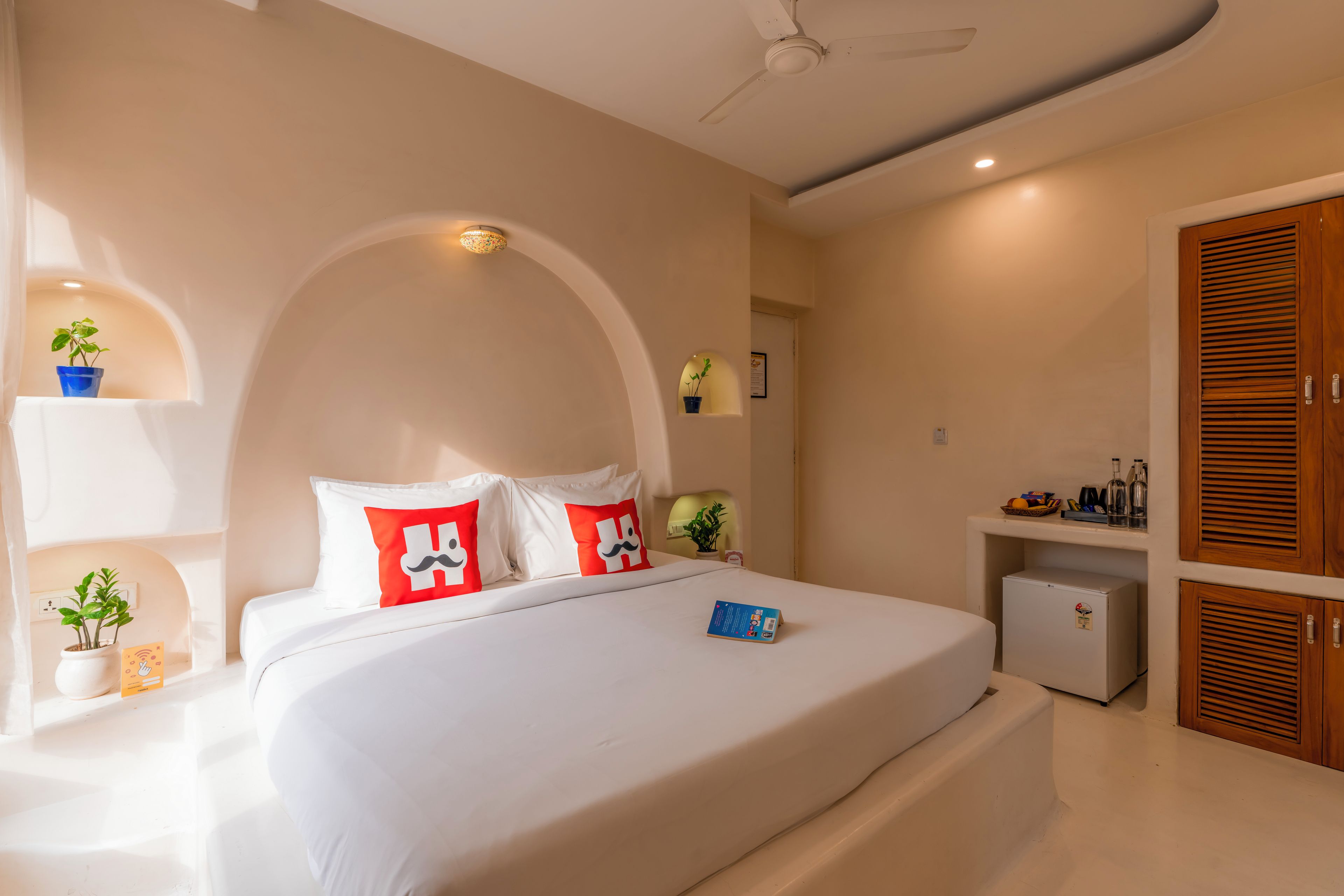 A hotel room with a bed, a ceiling fan, a small refrigerator, and a wooden wardrobe.