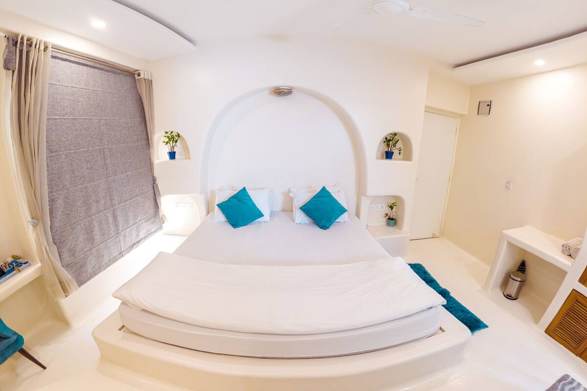 A modern bedroom with a curved bed, a large window with curtains, a small plant, and a bedside table.