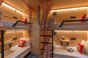 A train compartment with four bunk beds, each with a pillow and a blanket. There are wall-mounted fans and a small table with a book.