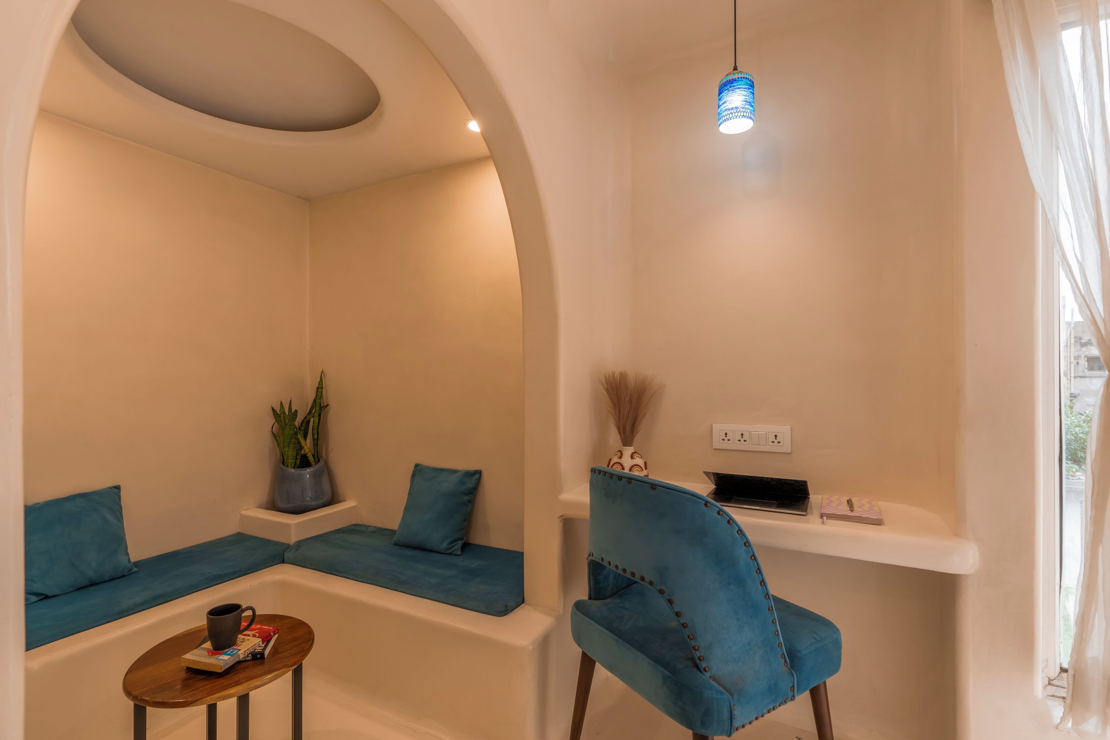 Um quarto com teto arredondado, um banco estofado azul, uma cadeira azul, uma pequena mesa com uma xícara e livros, e um lustre pendurado.