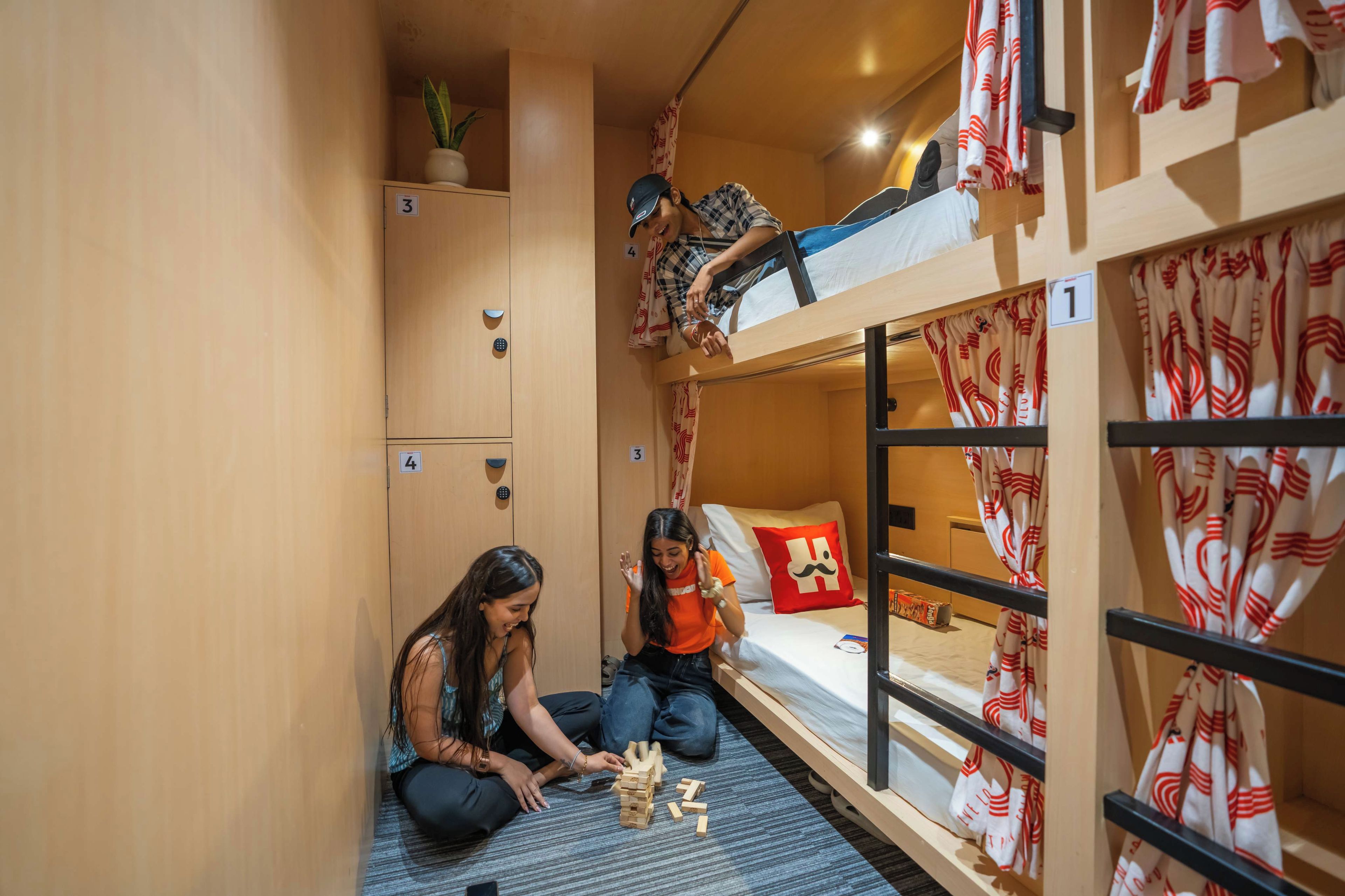 A bunk bed room with a game of Jenga. There are two people playing, a small plant on a shelf, and a suitcase on the floor.