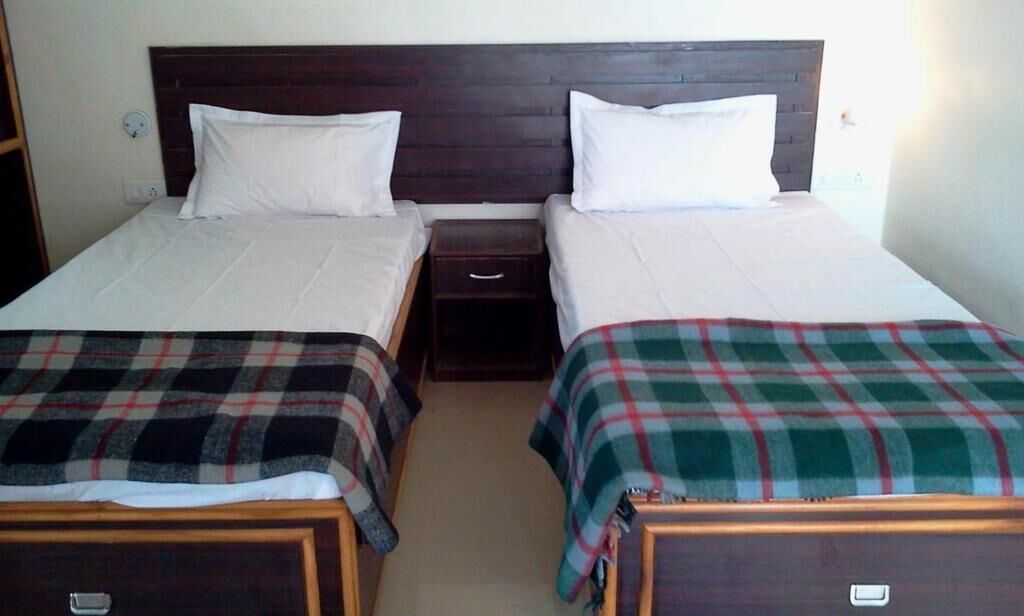 Una habitación doble con dos camas individuales, cada una con cabecero de madera y ropa de cama blanca. Entre las camas hay una mesita de noche de madera con un cajón.