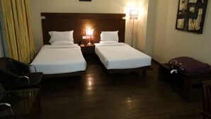 A hotel room with two single beds, a nightstand with a lamp, a wooden headboard, a framed picture on the wall, and a suitcase on a table. - Hotel Aane (Papumpare)