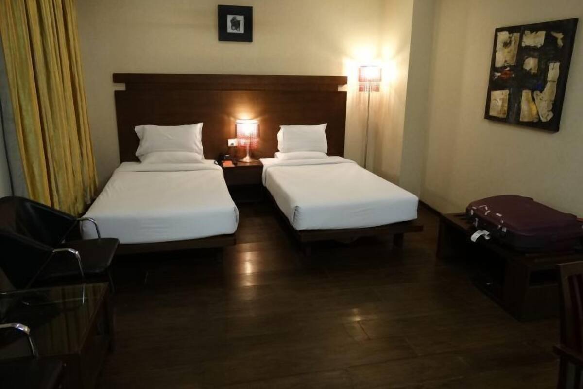 Hotellihuone, jossa on kaksi erillistä sänkyä, yöpöytä lampun kanssa, puinen pääty, seinällä oleva kehystetty kuva ja matkalaukku pöydällä.