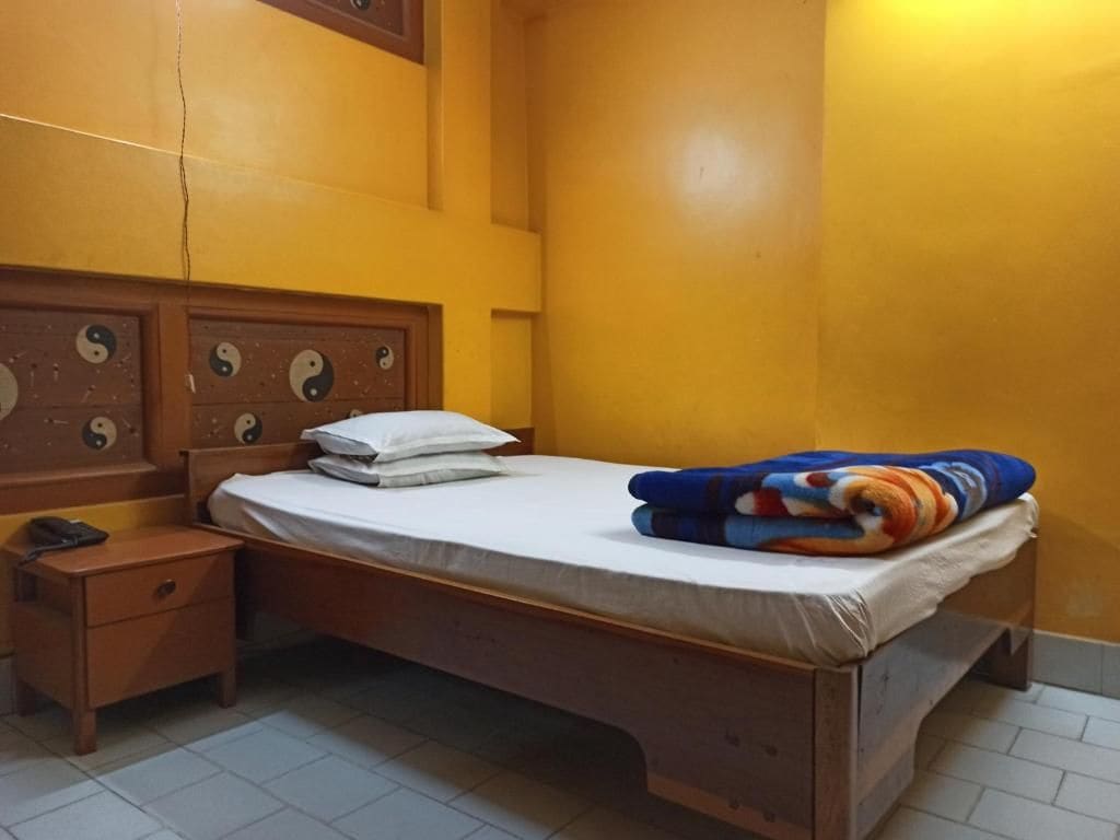 A single bed with a white mattress and a colorful blanket, a wooden nightstand with a phone, and a wall with a decorative panel.
