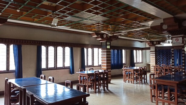 A spacious dining area with multiple wooden tables and chairs, large windows with blue curtains, and a grid-patterned ceiling. - Hotel Aane (Papumpare)
