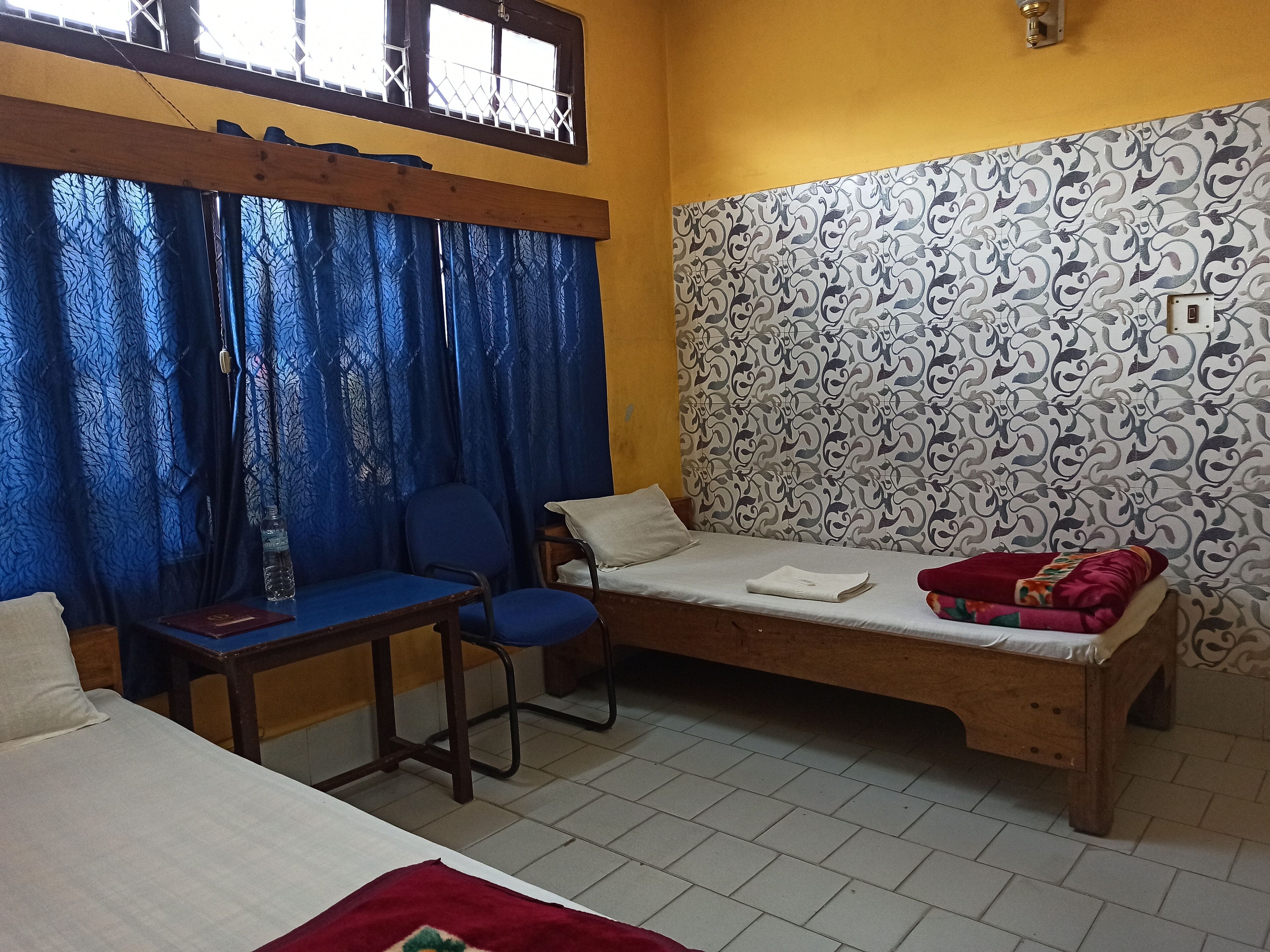 Une pièce comprenant un lit, un bureau et une chaise. Il y a une fenêtre avec une grille en métal et des rideaux bleus.