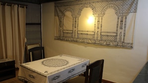 Ein Essbereich mit einem Tisch, bedeckt mit einer gemusterten Tischdecke, zwei Stühlen und einer Wandweberei mit einem architektonischen Motiv.