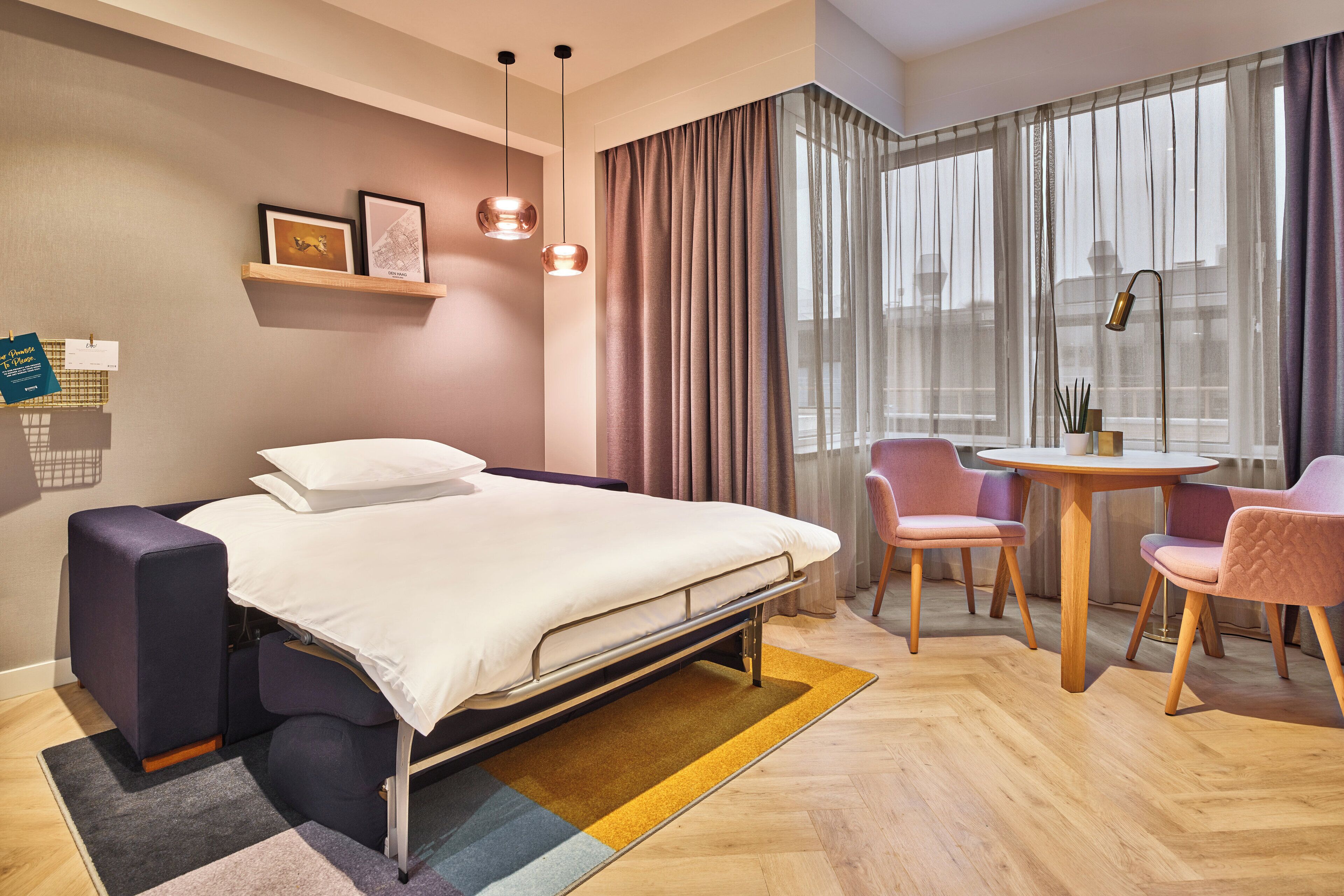 Una habitación de hotel moderna con una cama, un escritorio y sillas. Hay una ventana con cortinas, una lámpara y una estantería con fotografías.