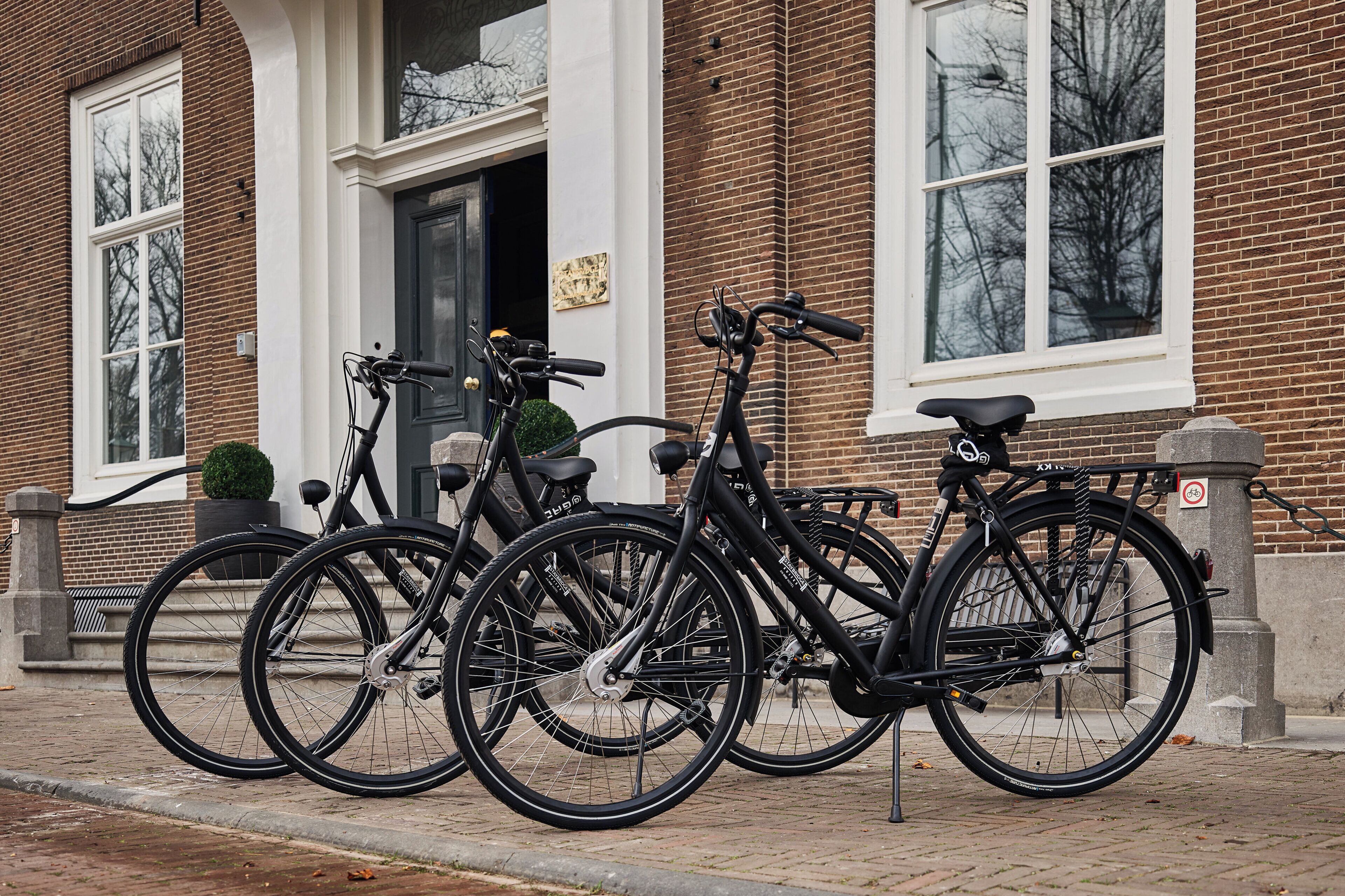 Trois vélos sont garés devant un bâtiment en briques, avec une porte et des fenêtres blanches.