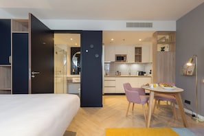 Une chambre d’hôtel moderne comprenant un lit, un coin repas avec deux chaises, une cuisine équipée d’un four à micro-ondes et une salle de bain avec des toilettes et un lavabo.