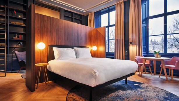 A modern hotel room with a large bed, a desk with two chairs, a bookshelf, and a view of the city through large windows.