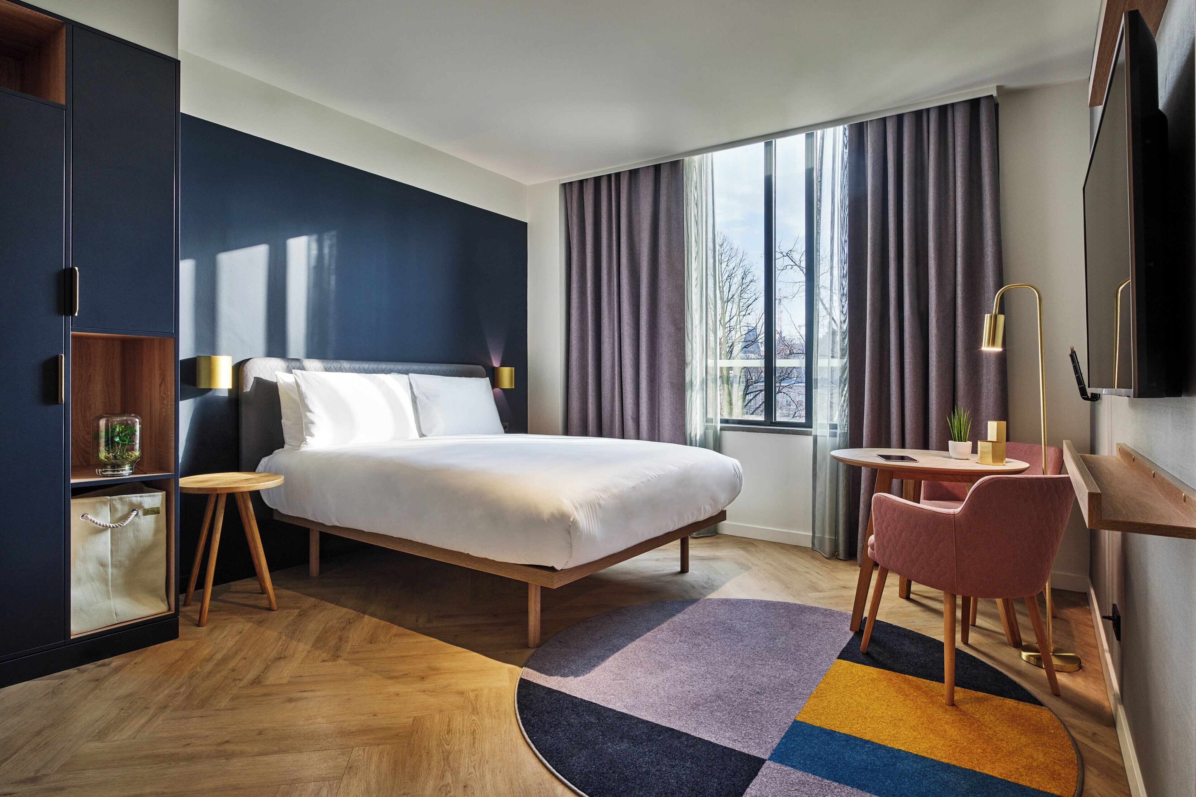 A modern hotel room with a large bed, a desk, a chair, a TV, and a window with curtains.