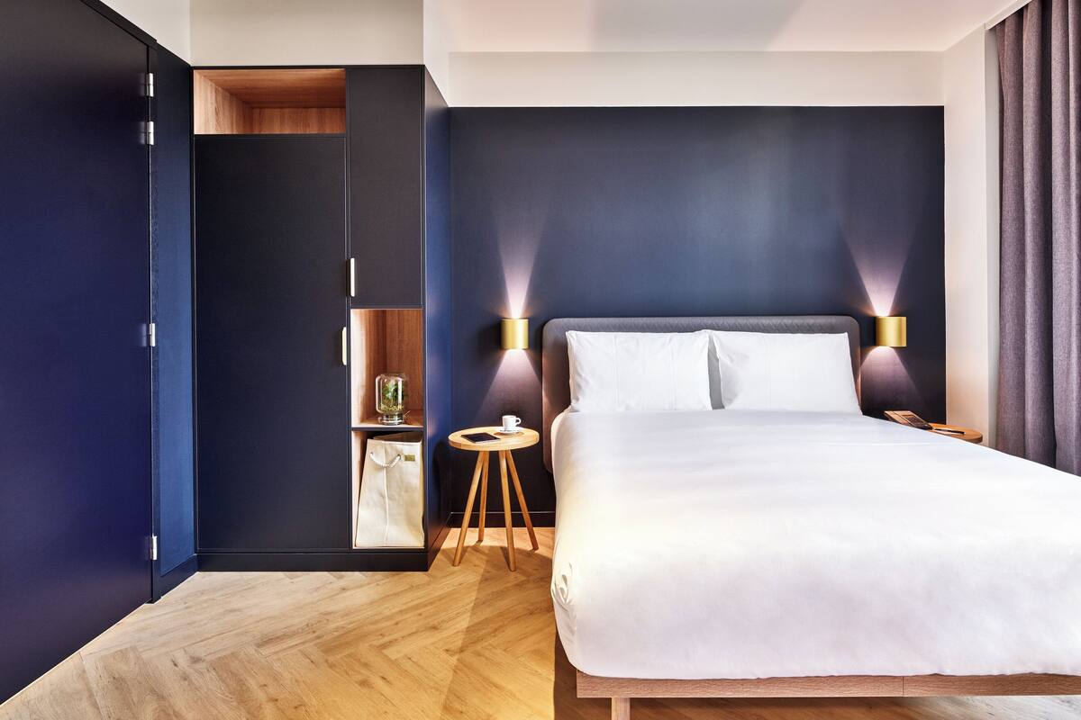 Een moderne hotelkamer met een groot bed, nachtlampjes, een klein houten nachtkastje en een garderobe met spiegel.
