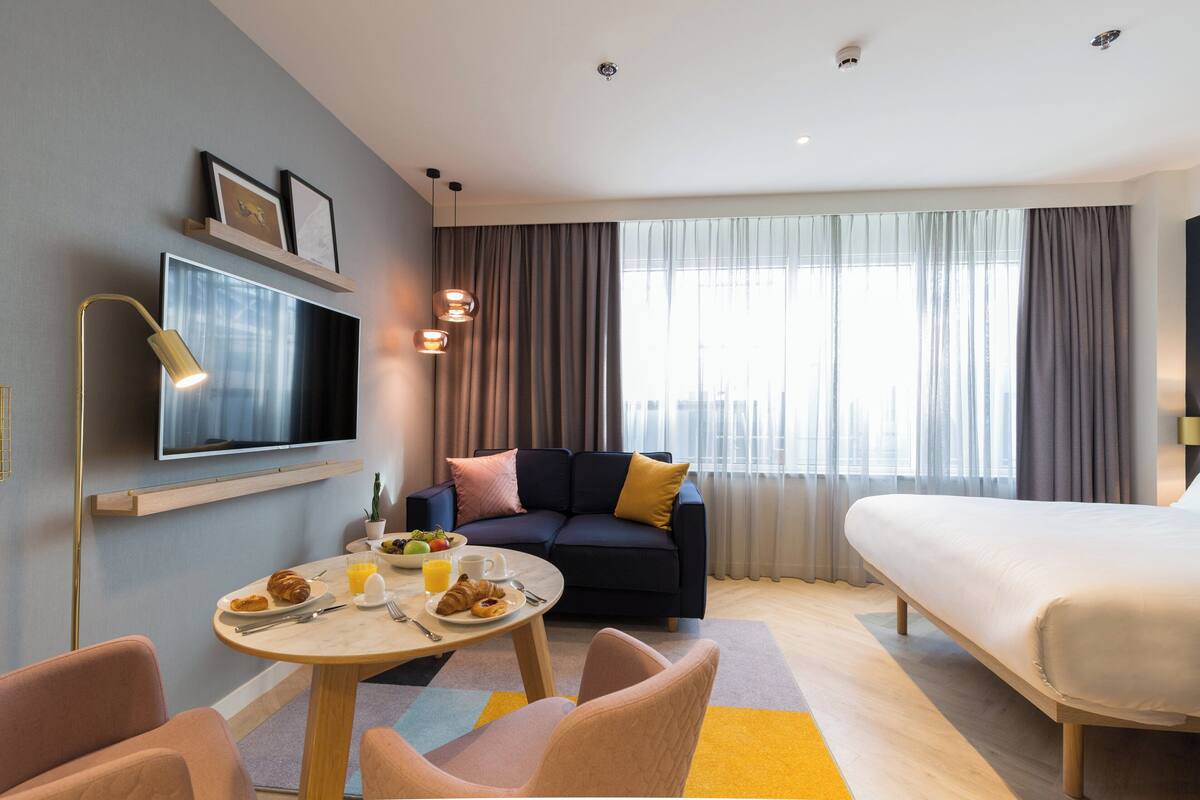 Een moderne hotelkamer met een bed, een sofa, een televisie, een eettafel met ontbijt en een groot raam met gordijnen.