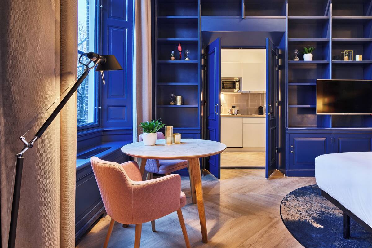 Een moderne kamer met een bed, een ronde tafel met een stoel, een televisie, en een keukenhoek zichtbaar door een geopende deur.
