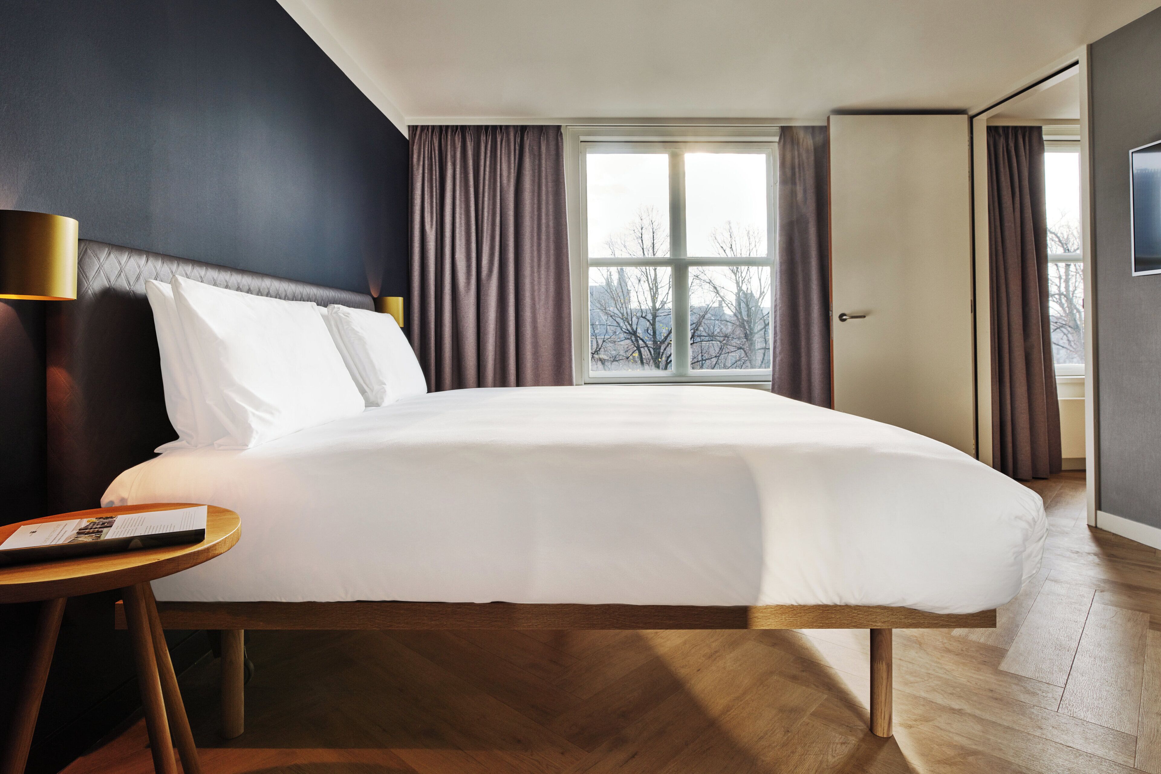 Quarto de hotel moderno com uma cama grande, uma mesa de cabeceira com abajur, e vista para o exterior pela janela.