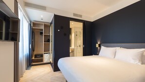 A modern hotel room with a large bed, a walk-in closet, a bathroom with a glass shower, and a view of the city.