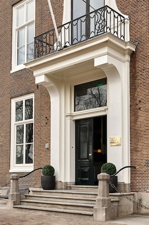 Een gebouw met twee verdiepingen, een zwarte voordeur, witte sierlijsten en een kleine tuin met twee planten in potten.