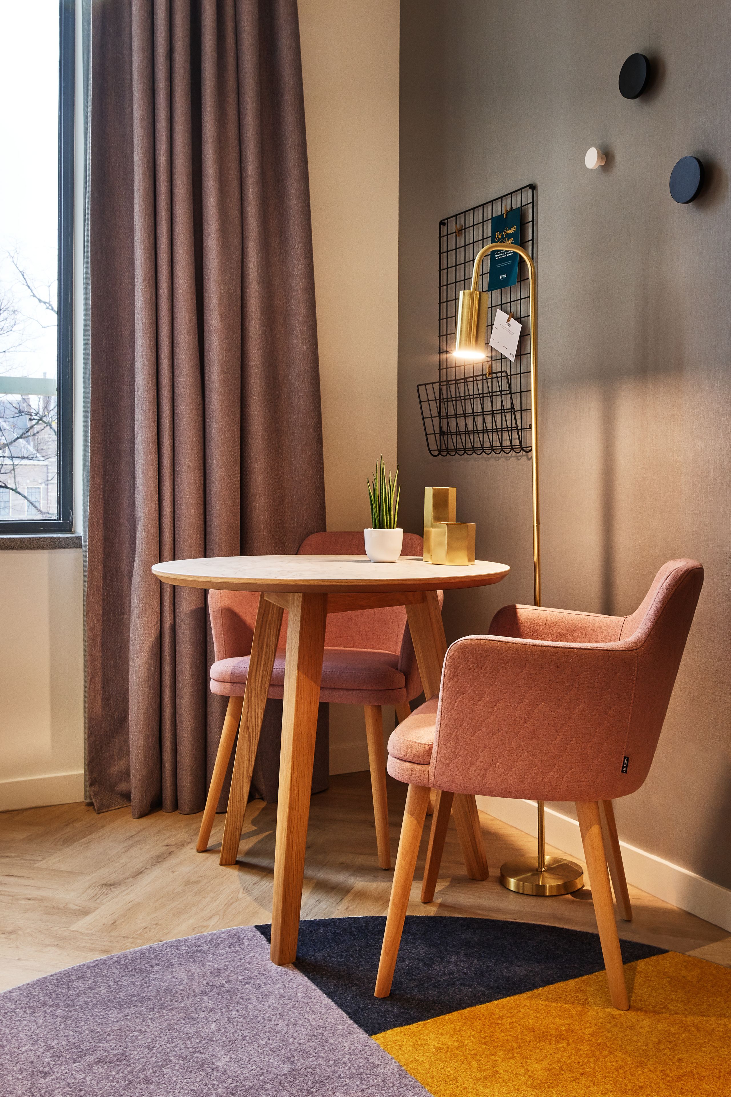A modern dining area with a round table, two pink chairs, a potted plant, and a floor lamp with a grid design.