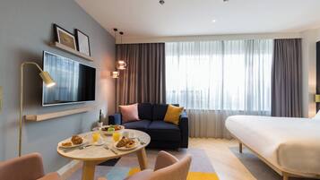 Una habitación de hotel con cama, sofá, mesa de comedor con desayuno, televisor y ventana con cortinas.