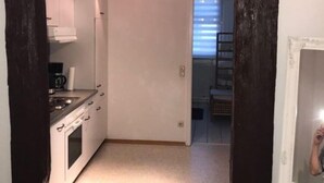 A narrow hallway leading to a kitchen with white cabinets and a stove. There is a window with blinds in the background. - Fachwerkhaus In Der Altstadt (Lueneburg)