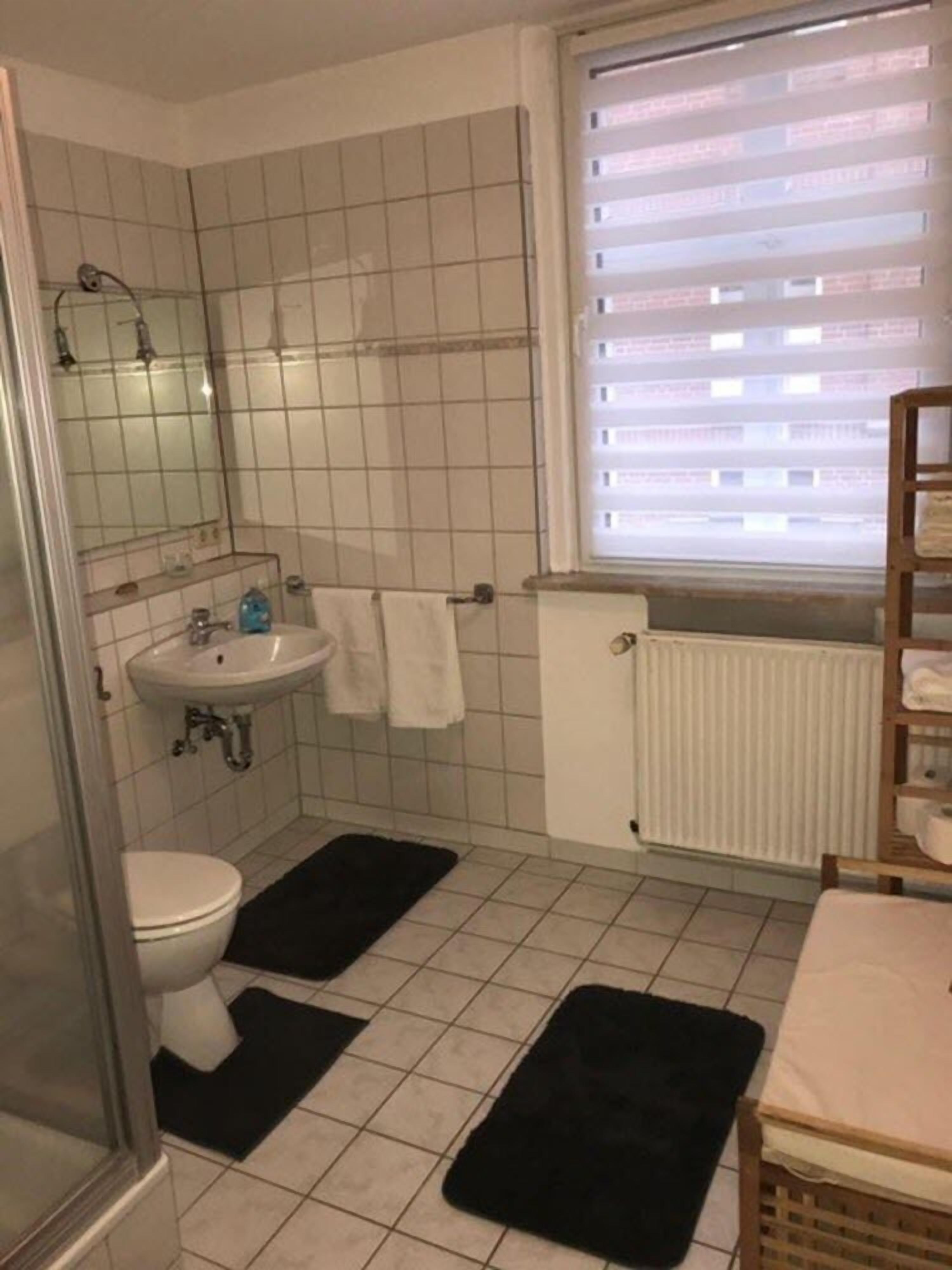 A bathroom with a white sink, mirror, and toilet. There are black rugs on the floor and a window with blinds.