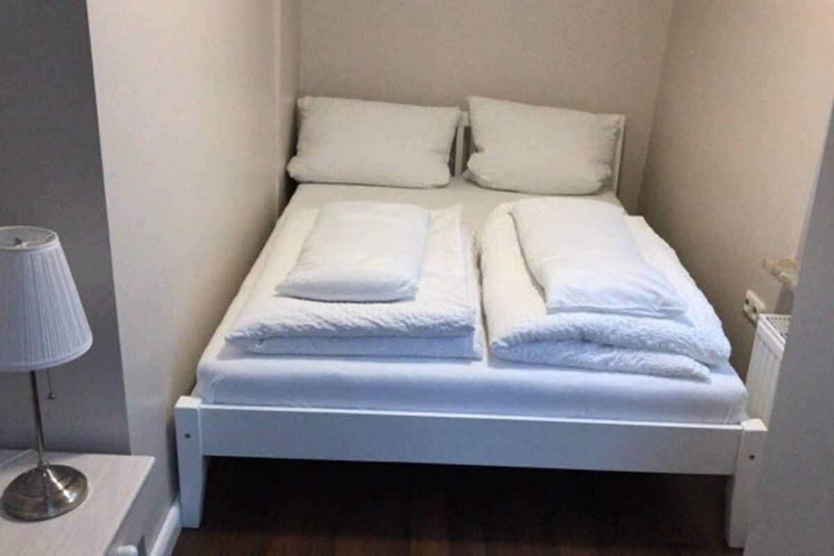 A small, neatly made bed with white linens and pillows, a bedside lamp, and a radiator.