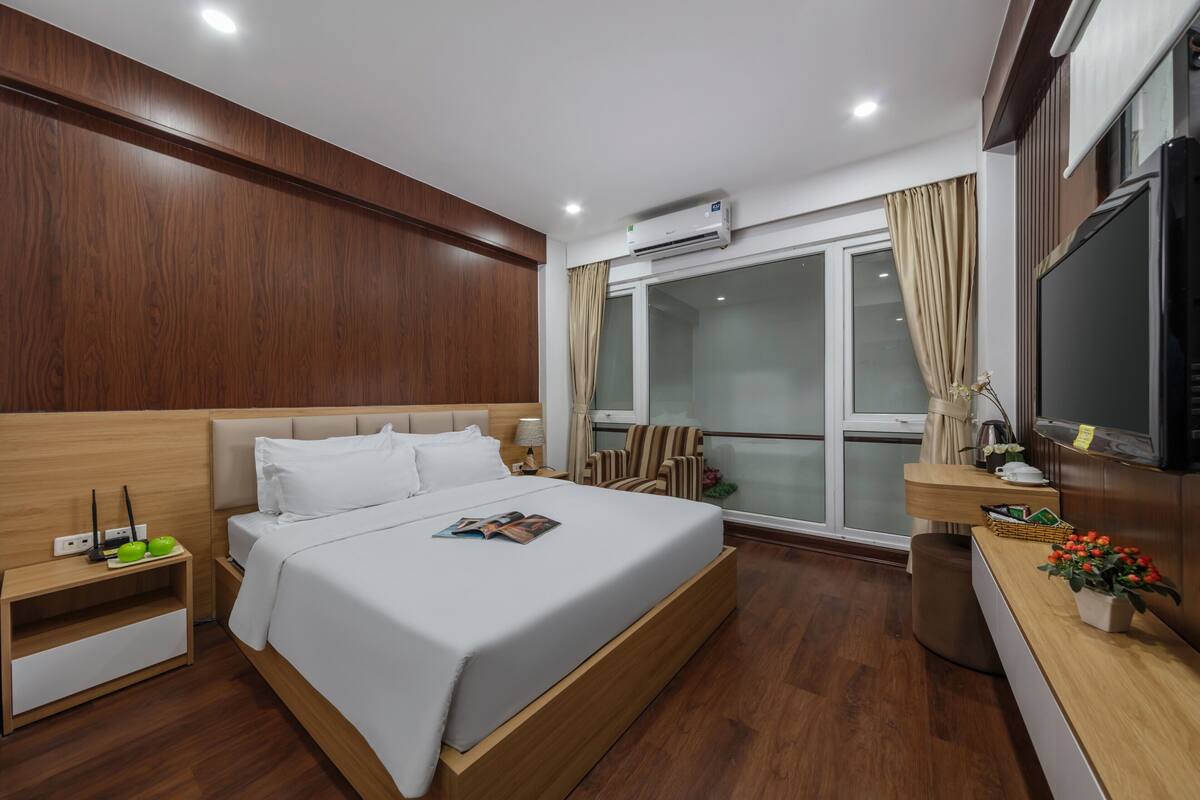 A modern hotel room with a wooden headboard, a large bed, a bedside table with a lamp, a TV, and a window with curtains.