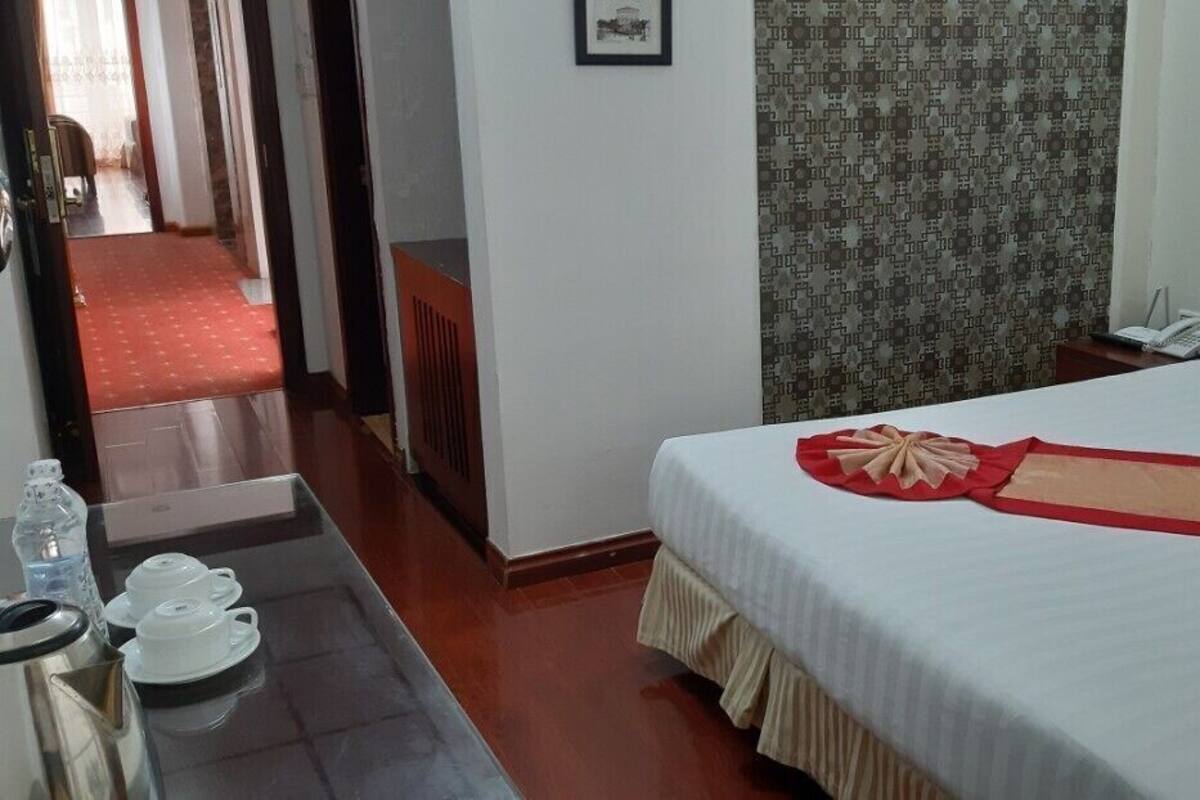 A hotel room with a bed, a bedside table with a phone, a red carpeted hallway, and a patterned wall.