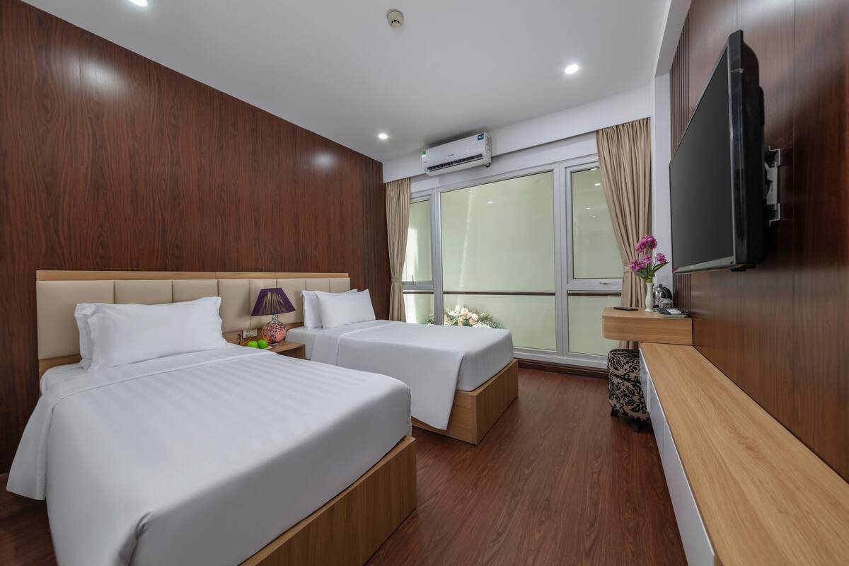 A hotel room with two beds, a TV, a window with curtains, and an air conditioning unit.