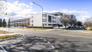 A modern building with a flat roof and large windows, situated at a street corner with a clear sky and a few clouds. - Accommodate Canberra - Dawes (Kingston)