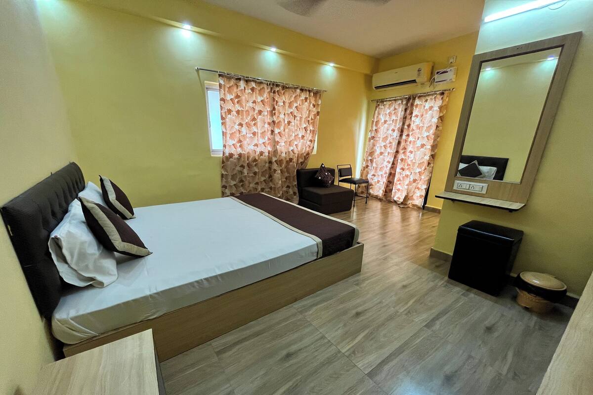 A hotel room with a bed, a small sofa, a chair, a mirror, and an air conditioning unit.