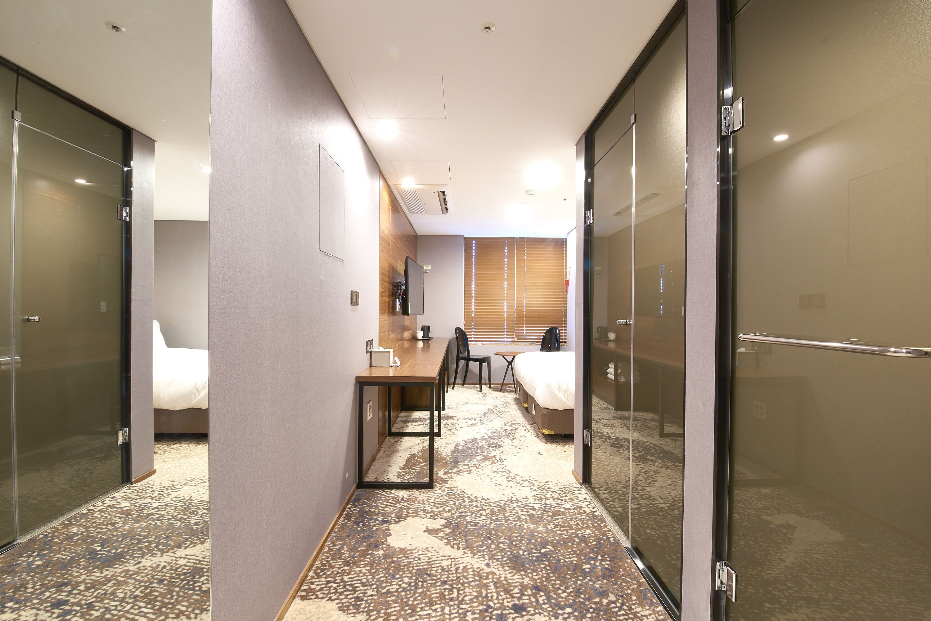 A hotel room with a bed, a desk, a chair, and a small table. There is a window with blinds, a door, and a glass partition.