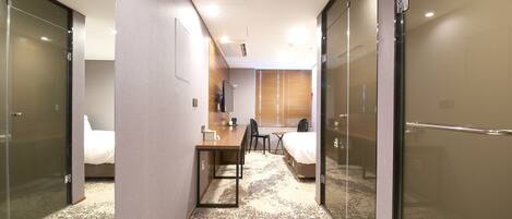 A hotel room with a bed, a desk, a chair, and a small table. There is a window with blinds, a door, and a glass partition.