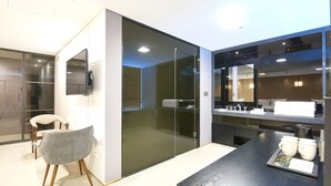 Ein modernes Hotelzimmer mit Glastrennwand, einem Holzstuhl, einer Kaffeestation und einem Flachbildfernseher.