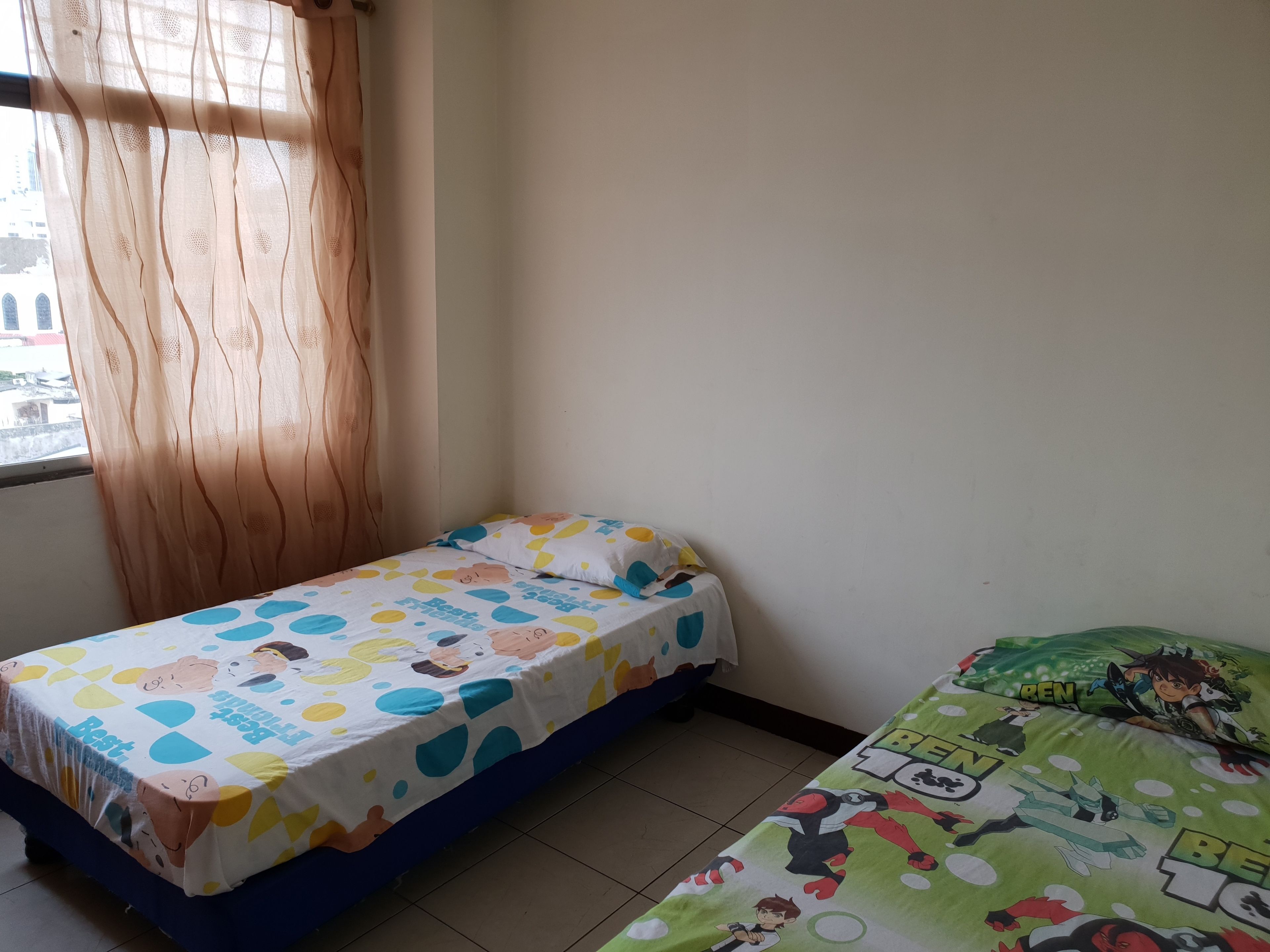 A small room with two beds, one with a colorful cartoon-themed bedspread and the other with a green superhero-themed bedspread.