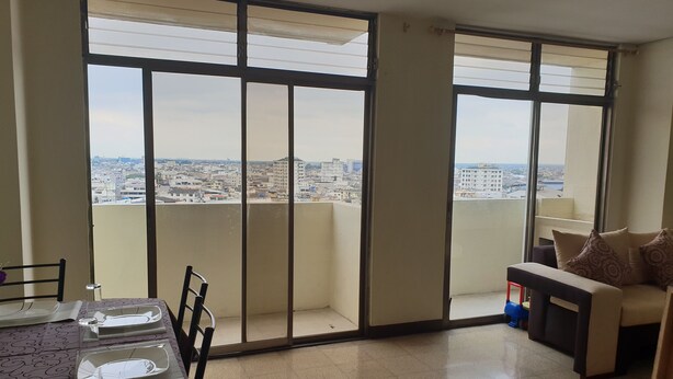 A dining area with a table set for two, a sofa, and a view of a cityscape through large windows. - Guayaquil In Love (Guayaquil)