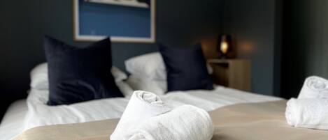 A neatly made bed with white linens and a beige throw blanket, pillows in dark and light shades, and a painting of a sunset over water on the wall.
