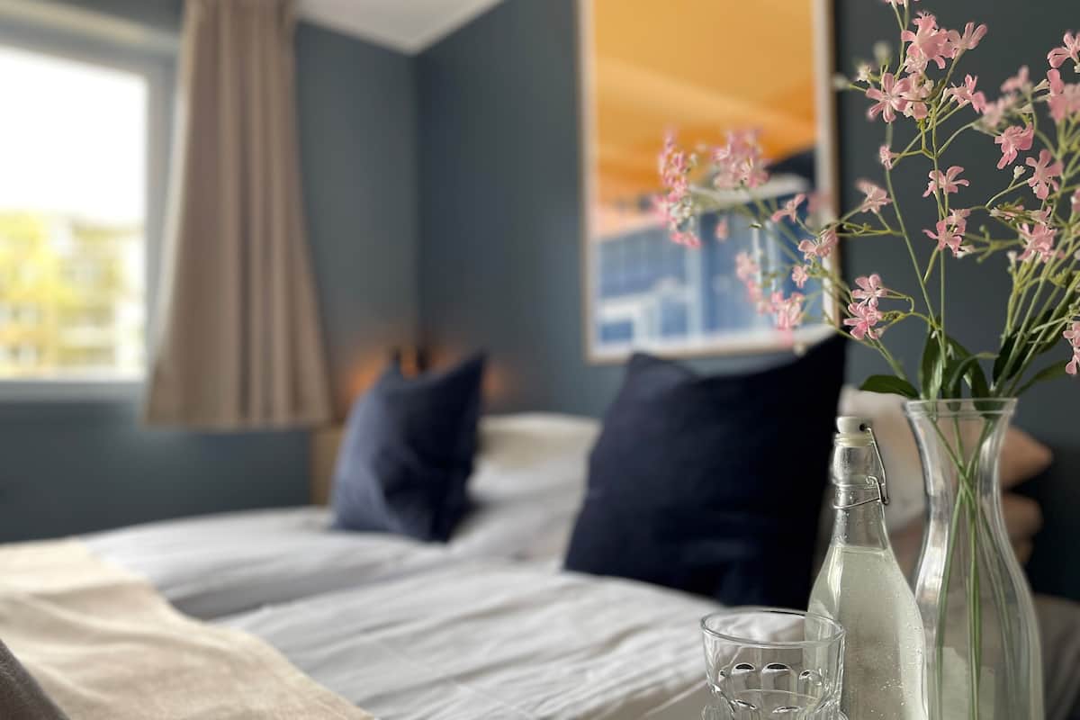 A neatly made bed with white linens, a dark blue pillow, and a beige throw blanket. There is a clear glass vase with pink flowers on the bedside table.