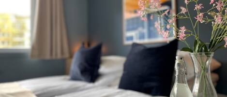Una cama bien tendida con sábanas blancas, una almohada azul oscuro y una manta color beige. Hay un florero de vidrio transparente con flores rosadas sobre la mesita de noche.