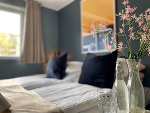 A neatly made bed with white linens, a dark blue pillow, and a beige throw blanket. There is a clear glass vase with pink flowers on the bedside table.