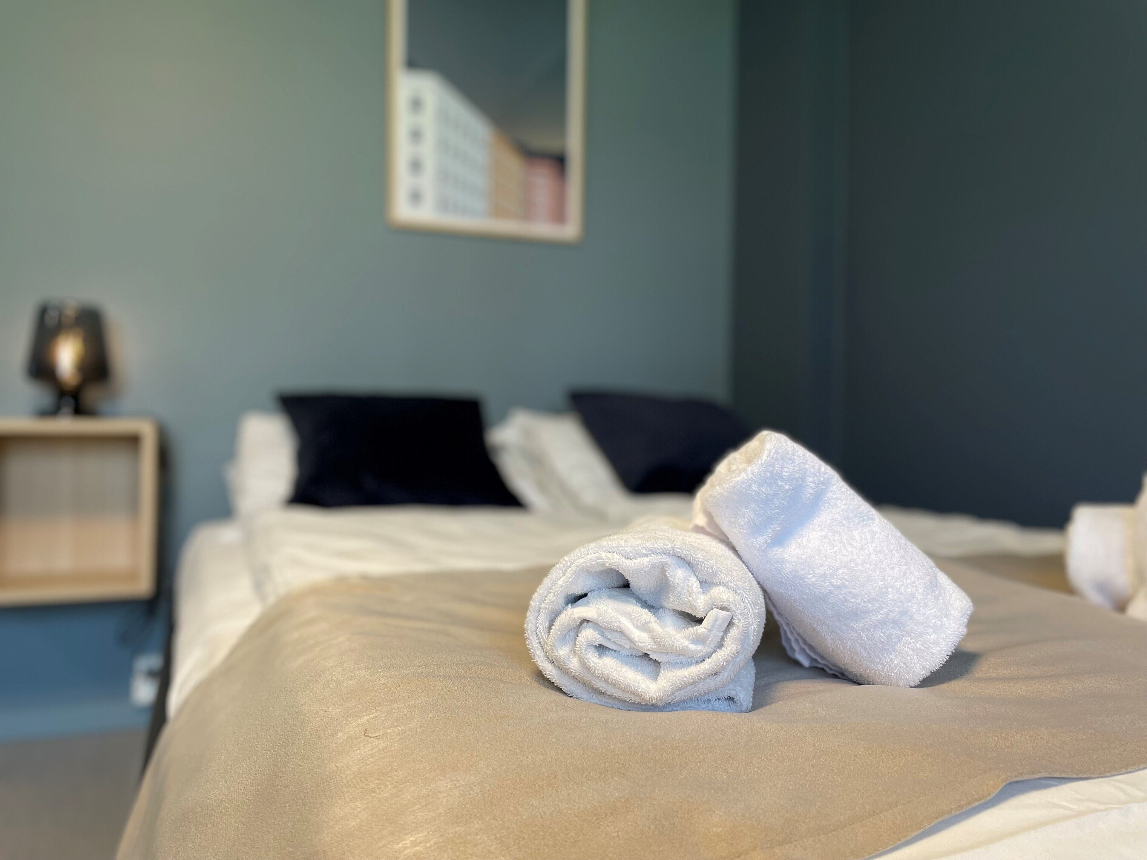 Una cama bien hecha con una colcha color beige y dos almohadas, una toalla blanca enrollada sobre la cama, y una mesita de noche con una lámpara y un adorno pequeño.