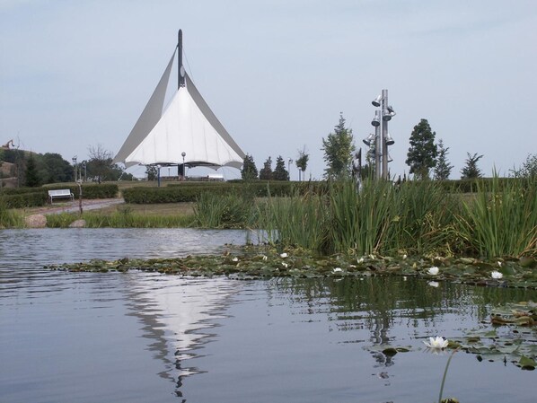 Una estructura arquitectónica moderna con techo blanco triangular, rodeada por una masa de agua con lirios y juncos, y un banco al frente.